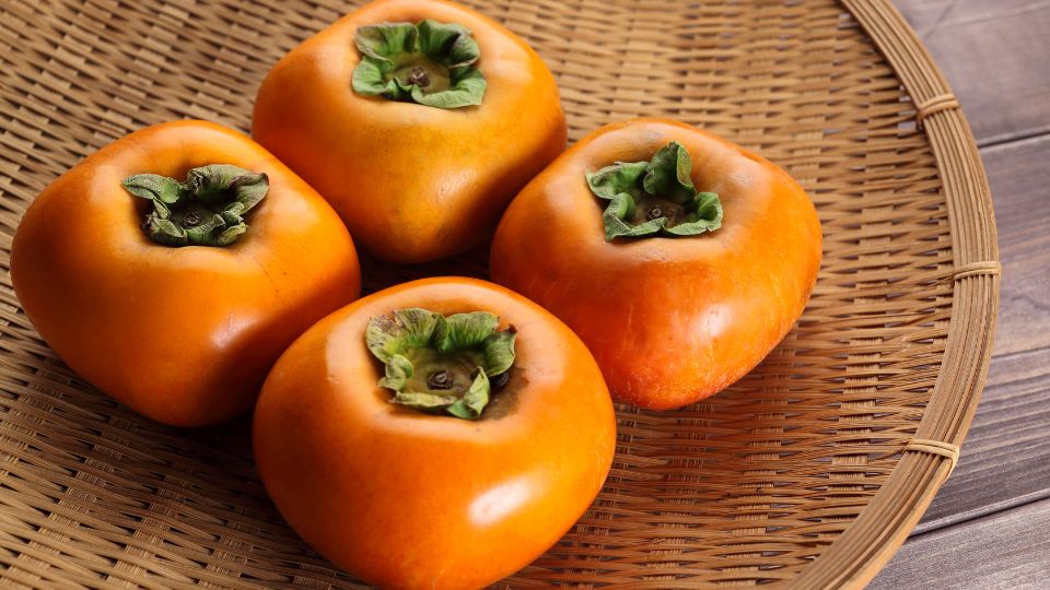 Vier reife Dattelpflaumen in einem Korb, leuchtend orange mit grünen Blättern.