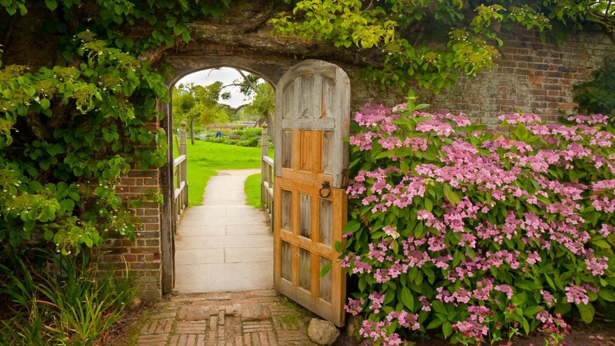 Ein offenes Holztor umgeben von blühenden Pflanzen und grünem Gras im Garten.