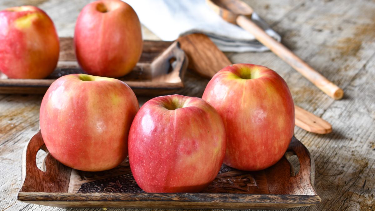 Drei frische Pink Lady-Äpfel auf einem Holztablett, leuchtend rot und gelb.