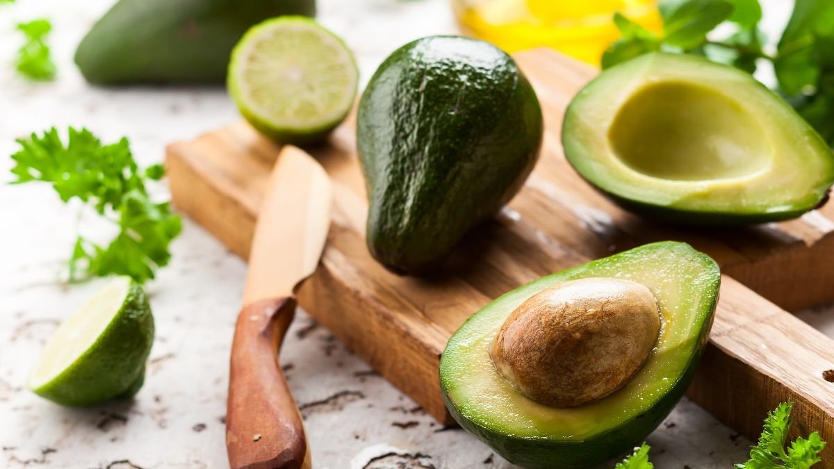 Eine aufgeschnittene Avocado mit dem Kern auf einem Holzbrett, umgeben von Limetten und Kräutern.