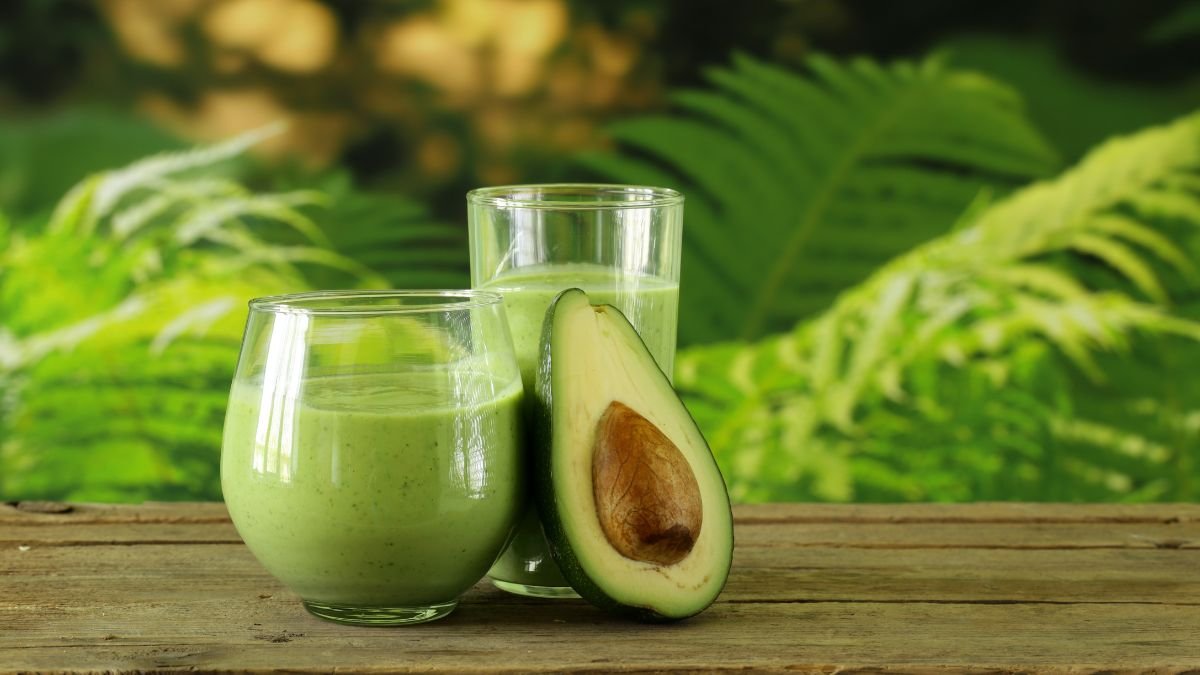 Zwei Gläser grüner Avocado-Smoothie neben einer halbierten Avocado auf einem Holztisch.