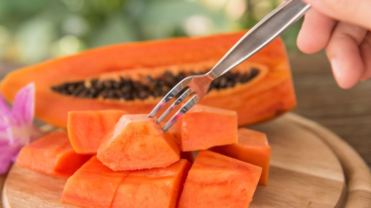 Eine Hand hält eine Gabel, um ein Stück frische Papaya zu essen, während die halbierte Frucht im Hintergrund sichtbar ist.